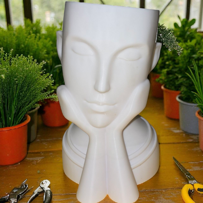 White ceramic sculpture of a face on a wooden table with gardening tools and plants in the background.